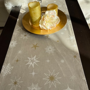 May include: A beige table runner with a gold and white star pattern. Two gold candles are on a gold charger plate with a white flower.