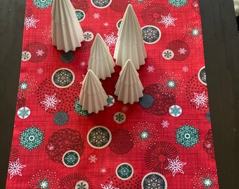 May include: A red table runner with a white snowflake and green circle pattern. Five white ceramic Christmas trees are arranged on the runner.