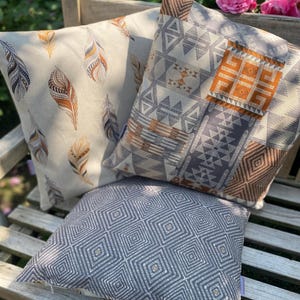 Puede incluir: Tres cojines cuadrados con patrones distintos. Uno presenta un fondo crema con plumas marrones y naranjas. Otro muestra un dise&ntilde;o geom&eacute;trico de patchwork en gris, blanco y naranja. El tercero tiene un patr&oacute;n de cuadr&iacute;cula de diamantes gris.