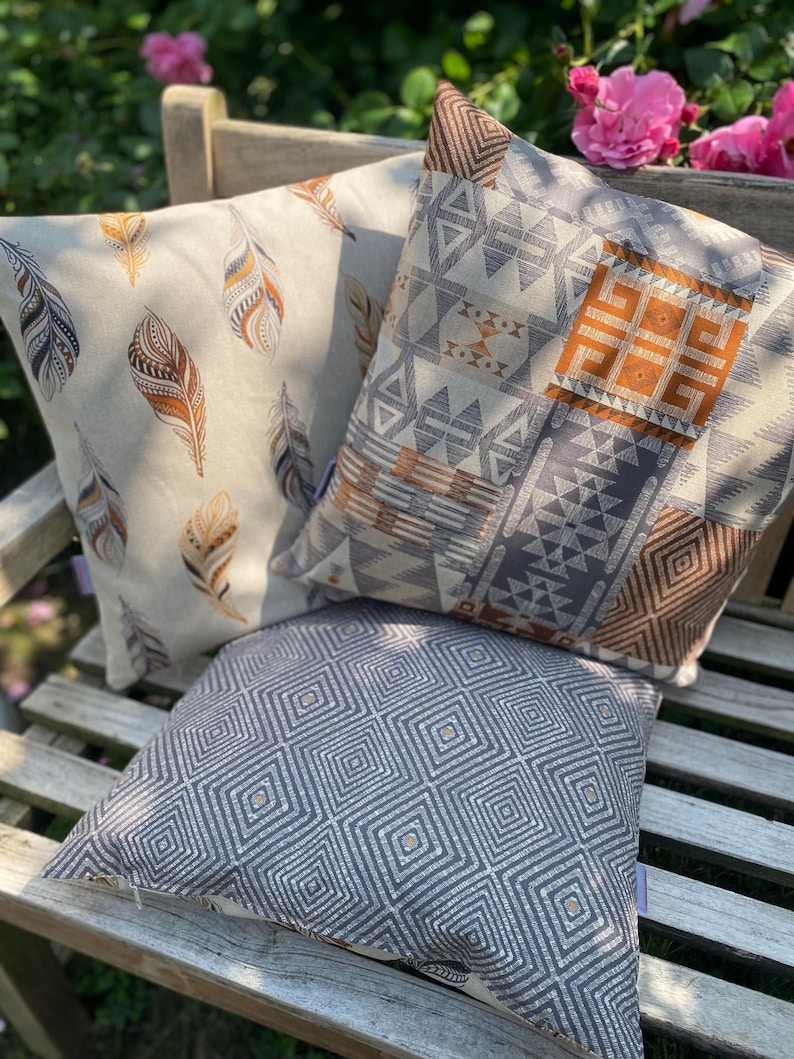 Puede incluir: Tres cojines cuadrados con patrones distintos. Uno presenta un fondo crema con plumas marrones y naranjas. Otro muestra un dise&ntilde;o geom&eacute;trico de patchwork en gris, blanco y naranja. El tercero tiene un patr&oacute;n de cuadr&iacute;cula de diamantes gris.