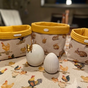 Op de afbeelding: Drie gele en witte stoffen opbergmanden met een wafelstructuur aan de bovenrand. De manden zijn gevuld met kleine knuffels en staan op een tafel met een wit tafelkleed met een patroon van gele kippen. Twee witte keramische eieren staan voor de manden.