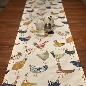 May include: White table runner featuring a repeating pattern of colourful chickens. The chickens are illustrated in a whimsical style with various colours and patterns.