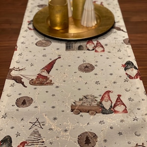Könnte beinhalten: Ein weißer Tischläufer mit einem festlichen Weihnachtsdesign mit Gnomen, Rentieren, Sternen und Bäumen. Der Läufer hat eine dezente Glitzer-Oberfläche und ist perfekt, um Ihrem Tisch einen Hauch von Weihnachtsfreude zu verleihen.