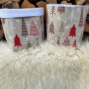 Pode incluir: Duas cestas de armazenamento de tecido com uma borda branca. As cestas são decoradas com um padrão de árvore de Natal em vermelho, marrom e branco sobre um fundo bege.
