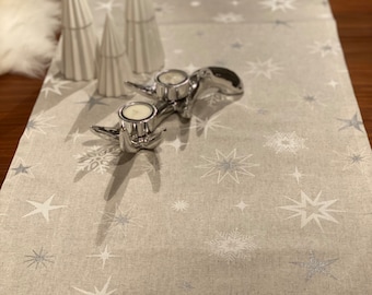 May include: A white table runner with a silver star and snowflake pattern. Three white cone-shaped decorations and two silver candle holders with candles are on the runner.