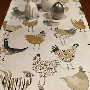 Könnte beinhalten: Ein weißer Tischläufer mit einem sich wiederholenden Muster aus braunen und schwarzen Hühnern. Die Hühner sind stilisiert und haben unterschiedliche Muster. Auf dem Tischläufer liegen drei dekorative Eier, zwei weiße und ein silbernes.