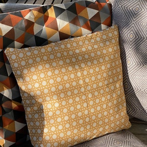 Puede incluir: Tres cojines cuadrados con patrones distintos. Un coj&iacute;n tiene un estampado de tejido de mimbre en color amarillo mostaza, otro muestra un patr&oacute;n geom&eacute;trico de tri&aacute;ngulos en naranja, gris y blanco, y el tercero tiene un dise&ntilde;o de rombos grises.