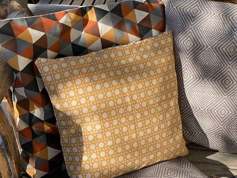 Puede incluir: Tres cojines cuadrados con patrones distintos. Un coj&iacute;n tiene un estampado de tejido de mimbre en color amarillo mostaza, otro muestra un patr&oacute;n geom&eacute;trico de tri&aacute;ngulos en naranja, gris y blanco, y el tercero tiene un dise&ntilde;o de rombos grises.