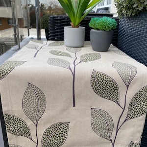 Puede incluir: Un camino de mesa beige con un patrón de hojas verdes. El camino de mesa está sobre una mesa de mimbre negra con dos plantas en macetas.