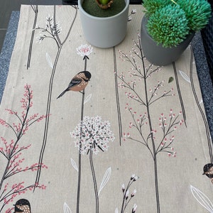 May include: A beige table runner with a floral pattern featuring small birds, white flowers, and pink berries. The runner is decorated with black stems and leaves.