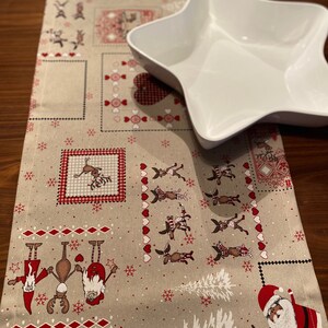 Könnte beinhalten: Ein beiger Tischläufer mit Weihnachtsmotiv mit roten und weißen Rentieren, Weihnachtsmann und Schneeflocken. Eine weiße sternförmige Servierplatte liegt auf dem Läufer.