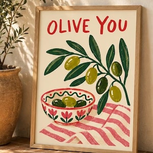 Op de afbeelding: Een ingelijste kunstdruk met de tekst OLIVE YOU in rode letters boven een illustratie van een groene olijftak en een witte kom met rode bloemmotieven gevuld met groene olijven, rustend op een roze en wit gestreepte doek.