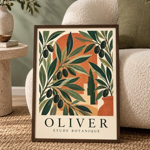 Op de afbeelding: Een ingelijste rechthoekige wandkunstprint met een botanische illustratie van olijftakken met donkergroene bladeren en zwarte olijven. De achtergrond toont een oranje boog en een terracotta pot. De tekst luidt OLIVER ÉTUDE BOTANIQUE.