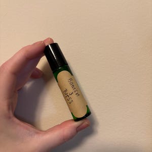 Puede incluir: Un frasco de aceite esencial en rollo de vidrio verde con tapa negra. El frasco tiene una etiqueta marrón con el texto escrito a mano "Banish & Bless". El frasco se sostiene sobre un fondo neutro.