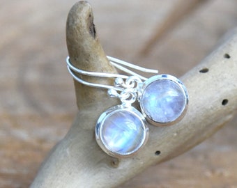 May include: A pair of silver dangle earrings with round moonstone gemstones. The earrings are on a wooden surface.