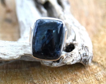 May include: A silver ring with a square black gemstone. The ring is sitting on a piece of driftwood.