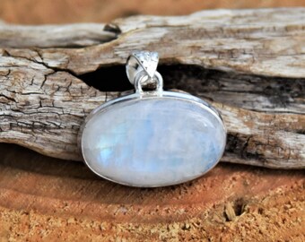 Puede incluir: Un colgante de plata con una piedra de luna ovalada blanca. El colgante está sobre una superficie de madera.