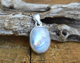Puede incluir: Un colgante de plata con una gran piedra de luna ovalada. La piedra de luna es iridiscente y tiene un color azul y blanco.