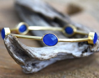 May include: A gold bangle bracelet with five blue oval-shaped gemstones. The bracelet is on a piece of driftwood.