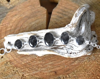 May include: A silver bracelet featuring oval-shaped black and gray stones. The stones are set in a silver chain with a toggle clasp. The bracelet is displayed on a piece of driftwood against a wooden background.
