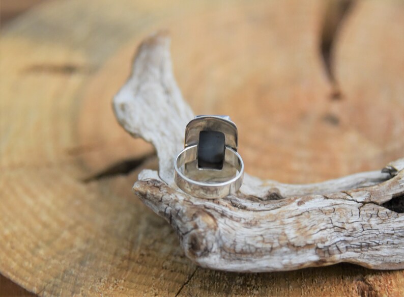 May include: A silver ring with a black stone set in a square bezel. The ring is on a piece of driftwood.
