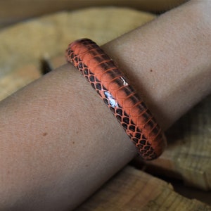 Puede incluir: Un brazalete de piel de serpiente de color naranja brillante y negro en una mu&ntilde;eca.