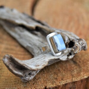 May include: A silver ring with a light blue rectangular stone set in a silver bezel. The ring is sitting on a piece of driftwood.