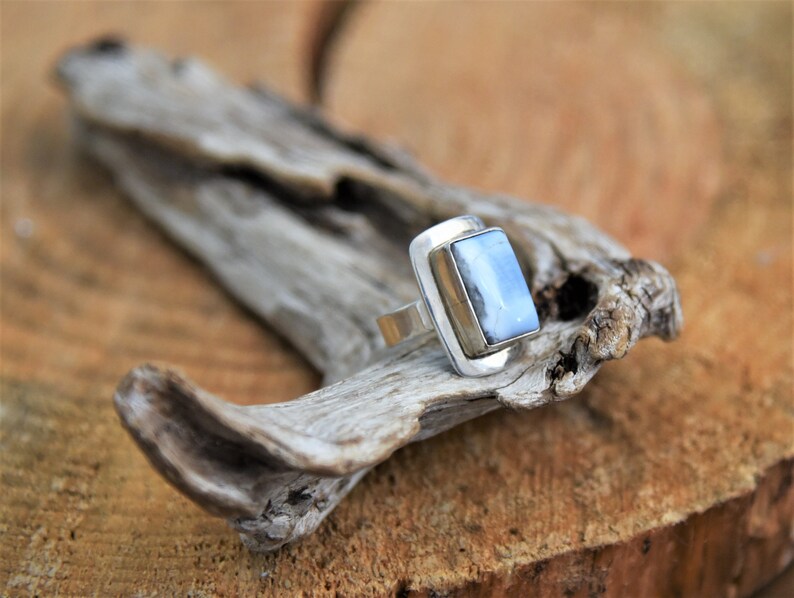 May include: A silver ring with a light blue rectangular stone set in a silver bezel. The ring is sitting on a piece of driftwood.