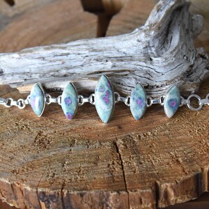 Könnte beinhalten: Ein silbernes Armband mit ovalen Edelsteinen. Die Edelsteine sind hellgrün mit rosa und roten Einschlüssen. Das Armband wird auf einer Holzoberfläche mit einem Stück Treibholz präsentiert.