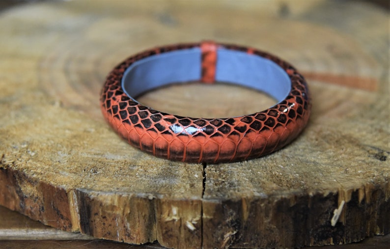 Puede incluir: Un brazalete de cuero con un patr&oacute;n de piel de serpiente roja y negra. El brazalete es liso y brillante.