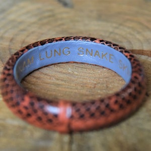 Puede incluir: Un brazalete de piel de serpiente marr&oacute;n y naranja con un interior azul. El brazalete est&aacute; grabado con el texto "ARM LUNG SNAKE SK".