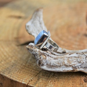 May include: A silver ring with a light blue gemstone set in a rectangular bezel. The ring is on a piece of driftwood.