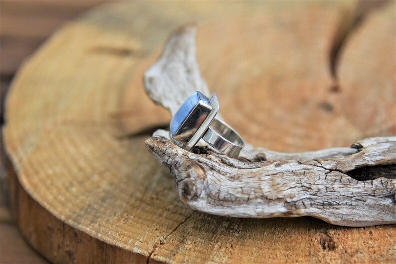 May include: A silver ring with a light blue gemstone set in a rectangular bezel. The ring is on a piece of driftwood.
