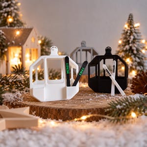 Op de afbeelding: Een witte en een zwarte skigondel als ornament, elk met een paar miniatuurski's aan de zijkant. De witte gondel heeft zwarte ski's met de groene tekst MERRY XMAS, terwijl de zwarte gondel witte ski's heeft met de tekst AHRRM XM.S.