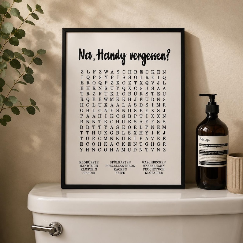 May include: A framed wall art print featuring a word search puzzle with the German text Na, Handy vergessen? at the top. The grid contains various letters and a list of words at the bottom, including Klob&uuml;rste, Handtuch, Klostein, and Pissoir.
