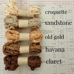 May include: Five bundles of crinkled ribbon arranged vertically, each secured with a tan paper band. The colors range from cream to dark brown, labeled as croquette, sandstone, old gold, havana, and claret on a vintage ledger paper background.