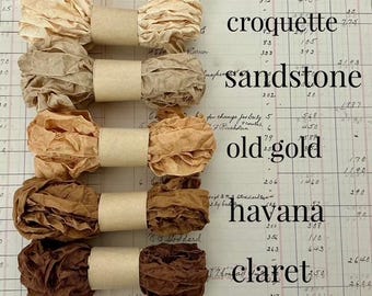 Könnte beinhalten: Fünf Bündel aus zerknittertem Band, vertikal angeordnet und jeweils mit einem beigen Papierstreifen fixiert. Die Farben reichen von Creme bis Dunkelbraun, beschriftet mit croquette, sandstone, old gold, havana und claret auf altem Papier.