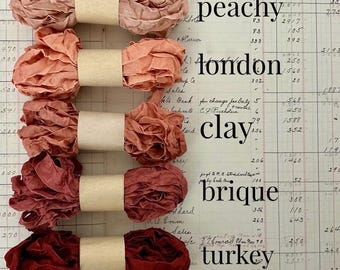 May include: Five spools of ruffled ribbon arranged vertically, each secured with a tan paper band. The colours range from light peach to dark red, labelled as peachy, london, clay, brique, and turkey next to each corresponding ribbon spool.