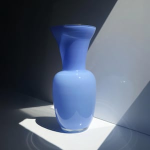 Könnte beinhalten: Eine hellblaue Glasvase mit glatter, opaker Oberfläche. Das Gefäß hat einen abgerundeten, bauchigen Boden, der sich zu einem schlanken, leicht ausgestellten Hals verjüngt, mit einer breiten, kreisförmigen Öffnung an der Oberseite in Pastellblau.