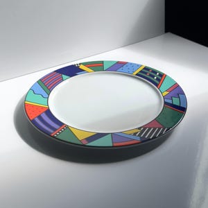 Könnte beinhalten: Ein runder Keramikteller mit weißer Mitte und einem breiten Rand, der mit einem bunten geometrischen Muster verziert ist. Der Rand zeigt verschiedene Formen in Blau-, Grün-, Gelb-, Rot- und Violetttönen, darunter Streifen, Punkte und Flächen.