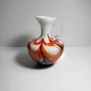 Könnte beinhalten: Eine weiße Glaskanne mit einem Wirbelmuster in Orange und Schwarz. Die Kanne hat einen durchsichtigen Glasgriff.