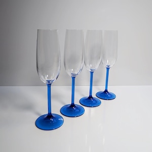 May include: Four clear champagne flutes with vibrant blue stems and bases are arranged in a row. The glasses are empty, showcasing their elegant design. The blue color provides a striking contrast against the clear glass.