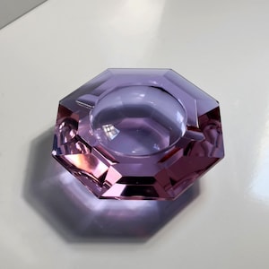 May include: An octagonal ashtray in shades of purple. The ashtray has a central circular depression and is made of a transparent material, reflecting light. The design features sharp angles and a polished finish.