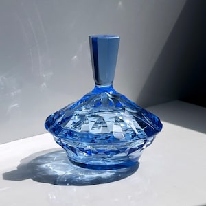 Könnte beinhalten: Ein facettierter, blauer Glasbehälter mit passendem Deckel. Der Behälter hat ein geometrisches Design und wird von Sonnenlicht beleuchtet, wodurch Schatten entstehen. Der Deckel ist ein dunklerer Blauton und hat eine zylindrische Form. Der Behälter dient wahrscheinlich zur Aufbewahrung kleiner Gegenstände.