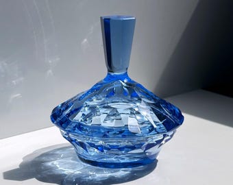 May include: A faceted, blue glass container with a matching lid. The container has a geometric design and is illuminated by sunlight, creating shadows. The lid is a darker shade of blue and has a cylindrical shape. The container is likely for storing small items.