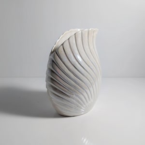 Könnte beinhalten: Eine perlmuttweiße Keramikvase mit einer einzigartigen, geschwungenen Form. Die Vase hat ein strukturiertes, geriffeltes Design, das sich um den Körper windet. Die Öffnung ist asymmetrisch, was ihren künstlerischen Reiz erhöht. Ein dekoratives Wohnaccessoire.
