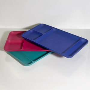 Könnte beinhalten: Drei farbige, unterteilte Tabletts in Blau, Türkis und Magenta. Jedes Tablett hat drei Fächer, mit einem größeren Abschnitt und zwei kleineren. Die Tabletts bestehen aus einem festen Material und sind übereinander gestapelt.