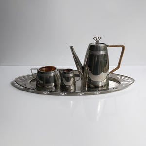 May include: A silver tea set on an oval tray. The set includes a teapot with a woven handle, a creamer, and a sugar bowl. The tray has a decorative edge. The teapot is approximately 8 inches tall.