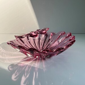 May include: A pink glass bowl with a scalloped edge and a decorative design. The bowl is made of clear glass with a pink tint.