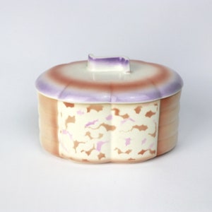 May include: A vintage ceramic trinket box with a floral design. The box is oval shaped and has a lid with a small handle. The box is decorated with a pink and white floral pattern on a cream background. The lid is decorated with a gradient of purple, pink, and brown.